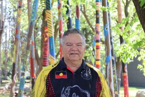 Man looks at camera and is standing in front of coloured poles. His shirt is black and yellow and has an Aboriginal flag on it.